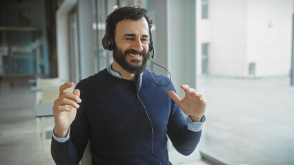 Handsome bearded man wearing a headset smiles engagingly in a modern office setting.