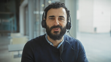 Handsome bearded man wearing headphones smiling in a bright modern office setting.