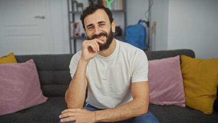 Fototapeta premium Smiling bearded man sitting comfortably at home on a grey couch with colorful cushions, portraying a relaxed lifestyle.