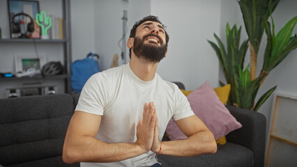 Bearded man in white shirt praying joyfully indoors with hands clasped, eyes closed, in a cozy...