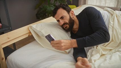 A bearded man lounges in bed while skeptically viewing his smartphone, reflecting the typical indoor life.
