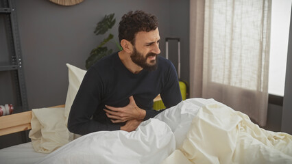 A bearded man in pain sitting on a bed indoors, clutching his stomach, evoking feelings of sickness or discomfort in a bedroom setting.