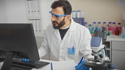 A bearded middle-aged man in a lab coat works at a computer in a laboratory, surrounded by...