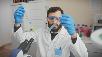 A middle-aged bearded man in lab goggles examines a test tube inside a modern laboratory.