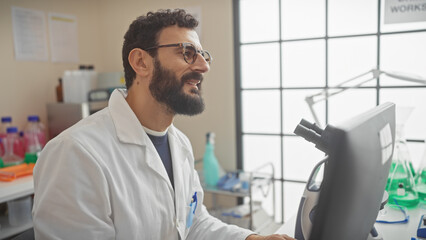 Smiling bearded scientist in white coat working at a computer in a bright laboratory.
