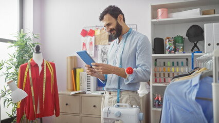 Bearded man using tablet in a bright tailor shop with mannequin, colorful threads, sewing machine,...