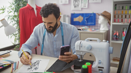 Fototapeta premium Handsome man tailoring in a modern atelier, using phone while sketching a fashion design.