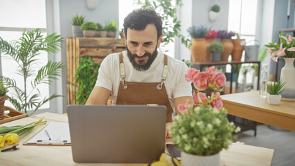 Bearded man using laptop in a vibrant flower shop with plants and apron