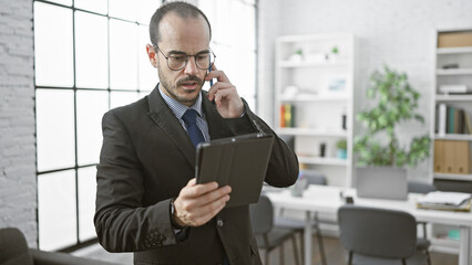 A professional man in a suit engaging in a business call while examining his tablet in a modern office interior.