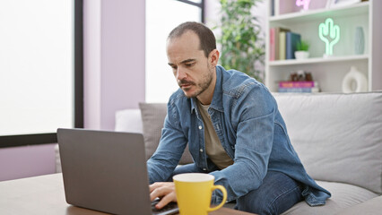 Hispanic bald man with beard focused on laptop in a modern living room with sofa and cactus decor.