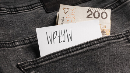  White card with a handwritten inscription "Wpływ", inserted into the pocket of gray pants jeasnow, next to Polish banknotes PLN (selective focus), translation: influence
