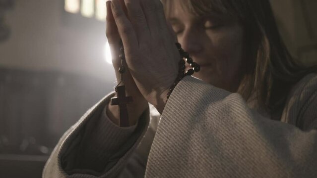 Religious woman holding beads with cross in folded arms while praying in church close up. Rosary in female Christian parishioner hands closeup. Religion symbol concept