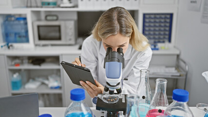 Blonde woman scientist using microscope in lab with digital tablet and chemicals