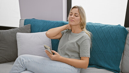 A young caucasian woman experiencing neck pain while holding a smartphone in a modern living room.