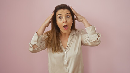 A surprised young hispanic woman with hands on her head against a pink background, portraying...