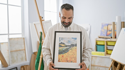 Handsome mature hispanic man holding artwork in bright studio interior.