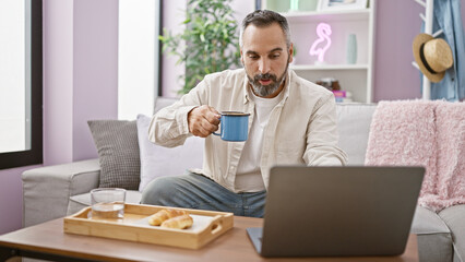 Mature hispanic man with beard working on laptop at home while holding a coffee cup on the sofa.