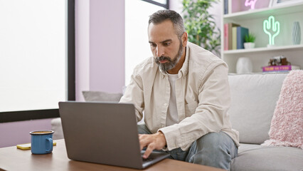 Handsome senior hispanic man with beard and grey hair using laptop on sofa indoors