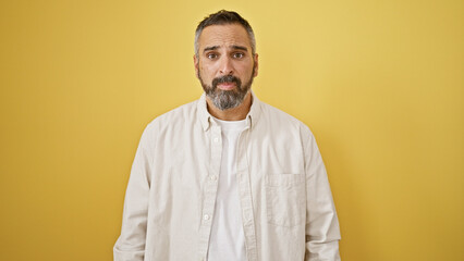 Mature hispanic man with beard and grey hair standing against a yellow wall, portraying a relaxed...