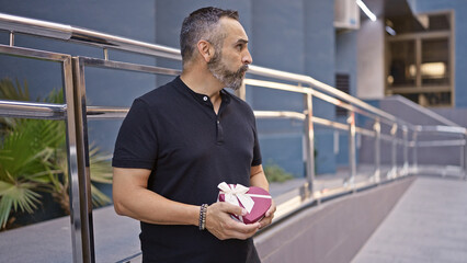 Mature hispanic man with beard and grey hair holding a gift on a city street.
