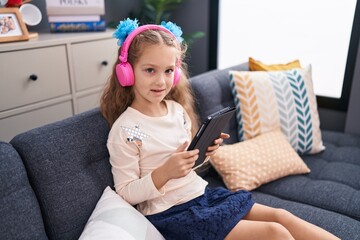 Adorable caucasian girl using touchpad and headphones sitting on sofa at home