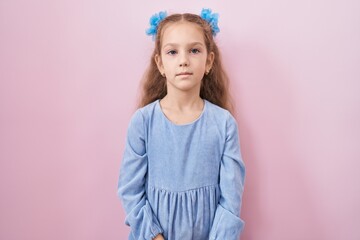 Young little girl standing over pink background with serious expression on face. simple and natural looking at the camera.