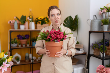 Young caucasian woman working at florist shop holding plant pot clueless and confused expression....