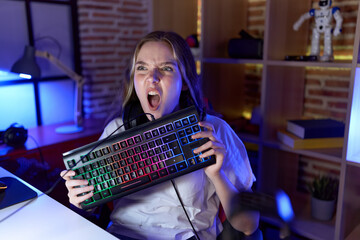 Young caucasian woman holding gamer keyboard angry and mad screaming frustrated and furious,...