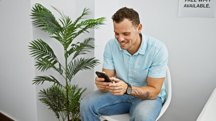 Smiling hispanic man with tattoo using smartphone in modern waiting room, conveying a casual and...