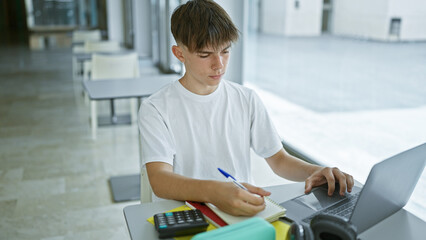 Caucasian teenage boy studying with laptop and books at university library table