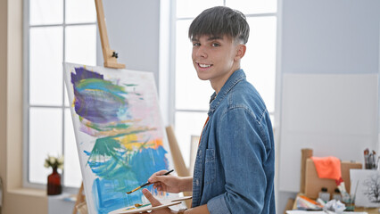 A young male artist painting on a canvas in a bright studio wearing a denim shirt.