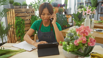 Obraz premium African american woman florist multitasking, talking on phone and using tablet in flower shop