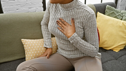 An adult woman with curly hair wearing a sweater sits indoors on a couch, hand on chest, in a cozy...