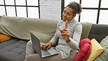 African american woman with curly hair gesturing no while using laptop on couch in home interior.