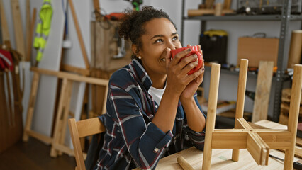 Smiling woman holding mug in woodworking studio with furniture projects around her.