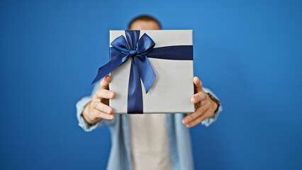A young hispanic man presents a wrapped gift with a blue ribbon against an isolated blue background.