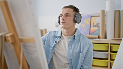 Pensive hispanic man with headphones stands in an art studio contemplating a canvas, embodying...