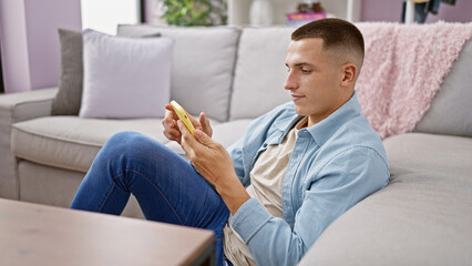 Handsome young man using smartphone on couch at home, reflecting modern lifestyle and connectivity.