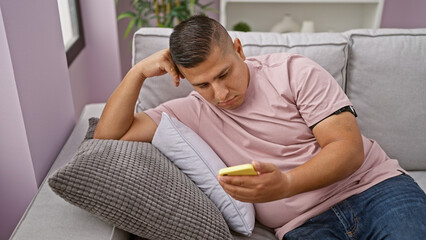 Obraz premium Handsome young latin man sitting upset on his living room sofa, engrossed in his smartphone texting woes, encapsulating indoor sadness at home