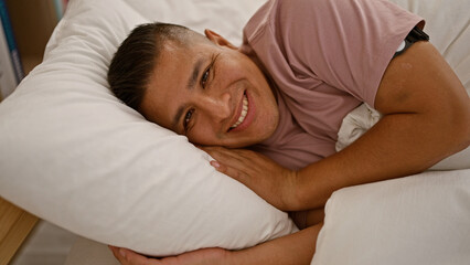 Confident young latin man blissfully relaxing, lying in bed, sporting a cheerful smile in the cozy ambiance of his comfortable bedroom.