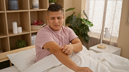 Handsome young latin man in pain, scratching itchy rash on arm while sitting on bedroom bed -...