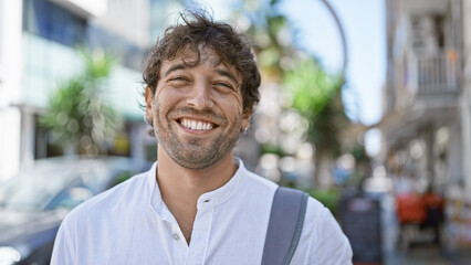 Smiling hispanic man with green eyes and a beard wearing a white shirt on a sunny city street.