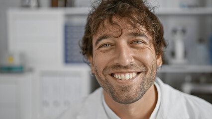 Smiling hispanic man with beard and green eyes wearing a lab coat in a hospital interior.