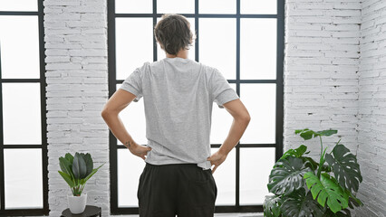 Back view of a man standing in a modern room with large windows and a white brick wall, conveying calmness and introspection.