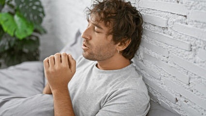 Relaxed hispanic man with beard and earring contemplating in a serene bedroom with white brick...