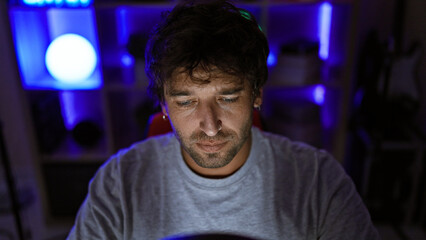 A focused hispanic man with a beard sits in a dark room at night, lit by ambient blue lights, giving off a gaming vibe.