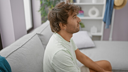 Thoughtful hispanic man with beard sits on living room sofa, showing profile in casual home setting