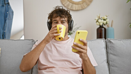 A relaxed hispanic man listens to music on headphones while holding a yellow mug and smartphone in...