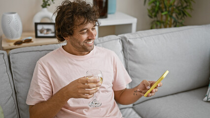 Smiling young hispanic man holding glass of wine and smartphone, sitting on couch indoors