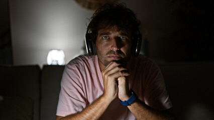 Thoughtful hispanic man with headphones in a dimly lit living room, contemplating